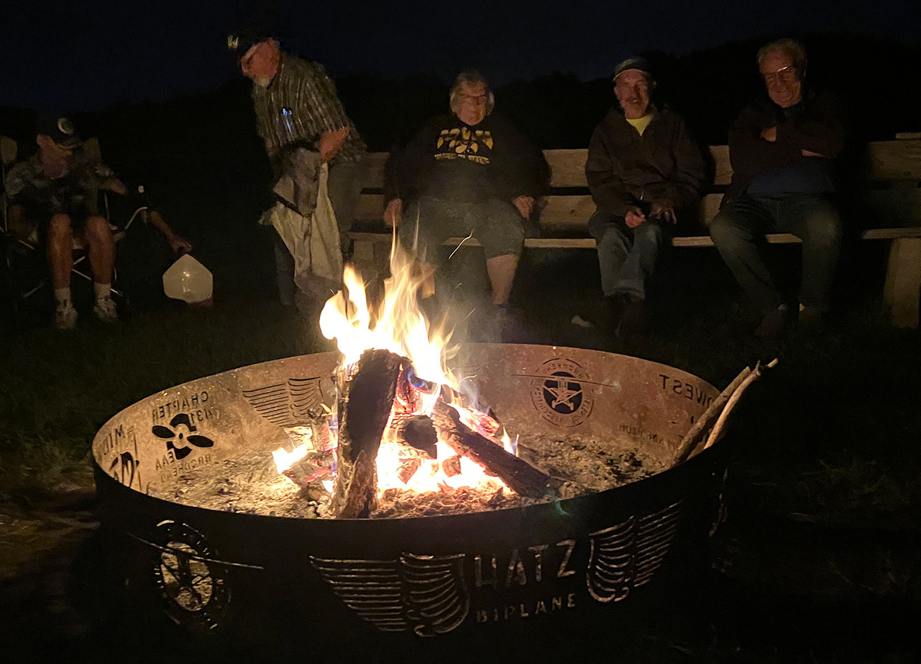 pilots gathered around a metal firepit at night. firepit has airplane motif cut out of sides.