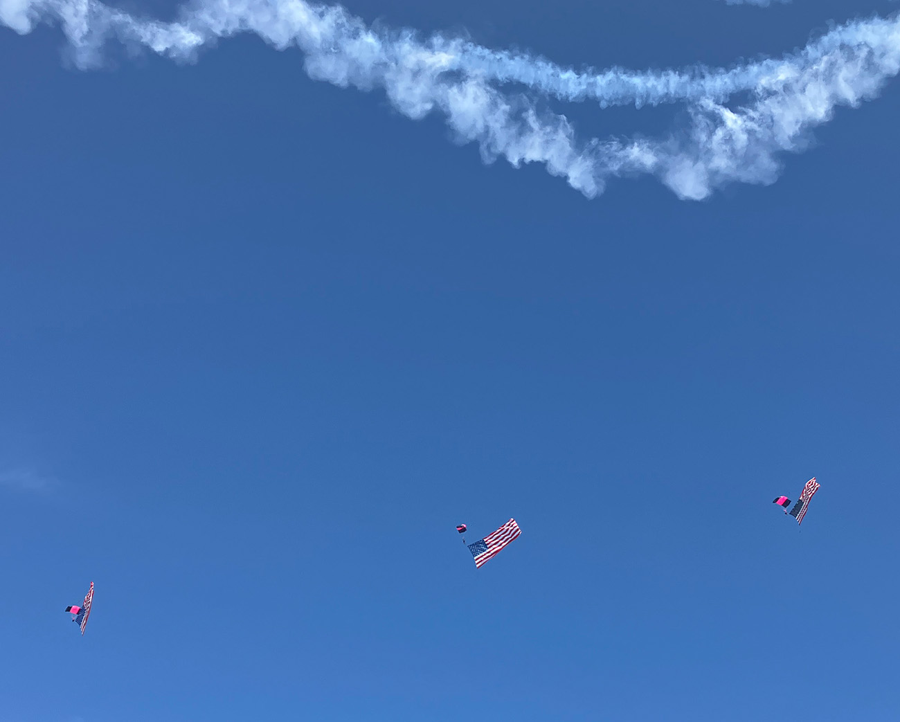 trail from airplane against blue sky, with american-flag carrying skydivers below it