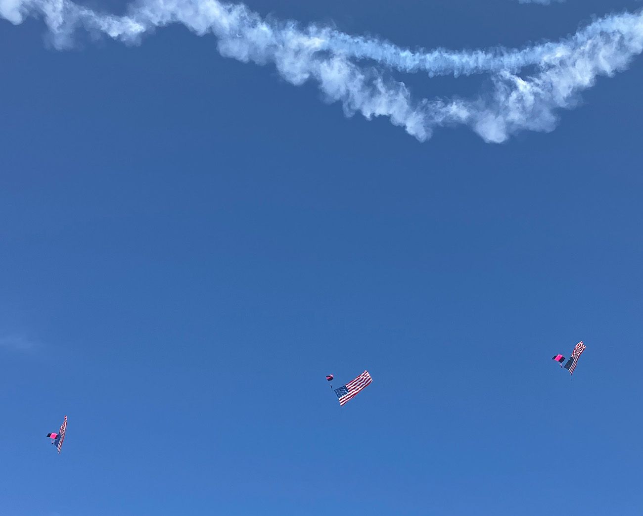 trail from airplane against blue sky, with american-flag carrying skydivers below it