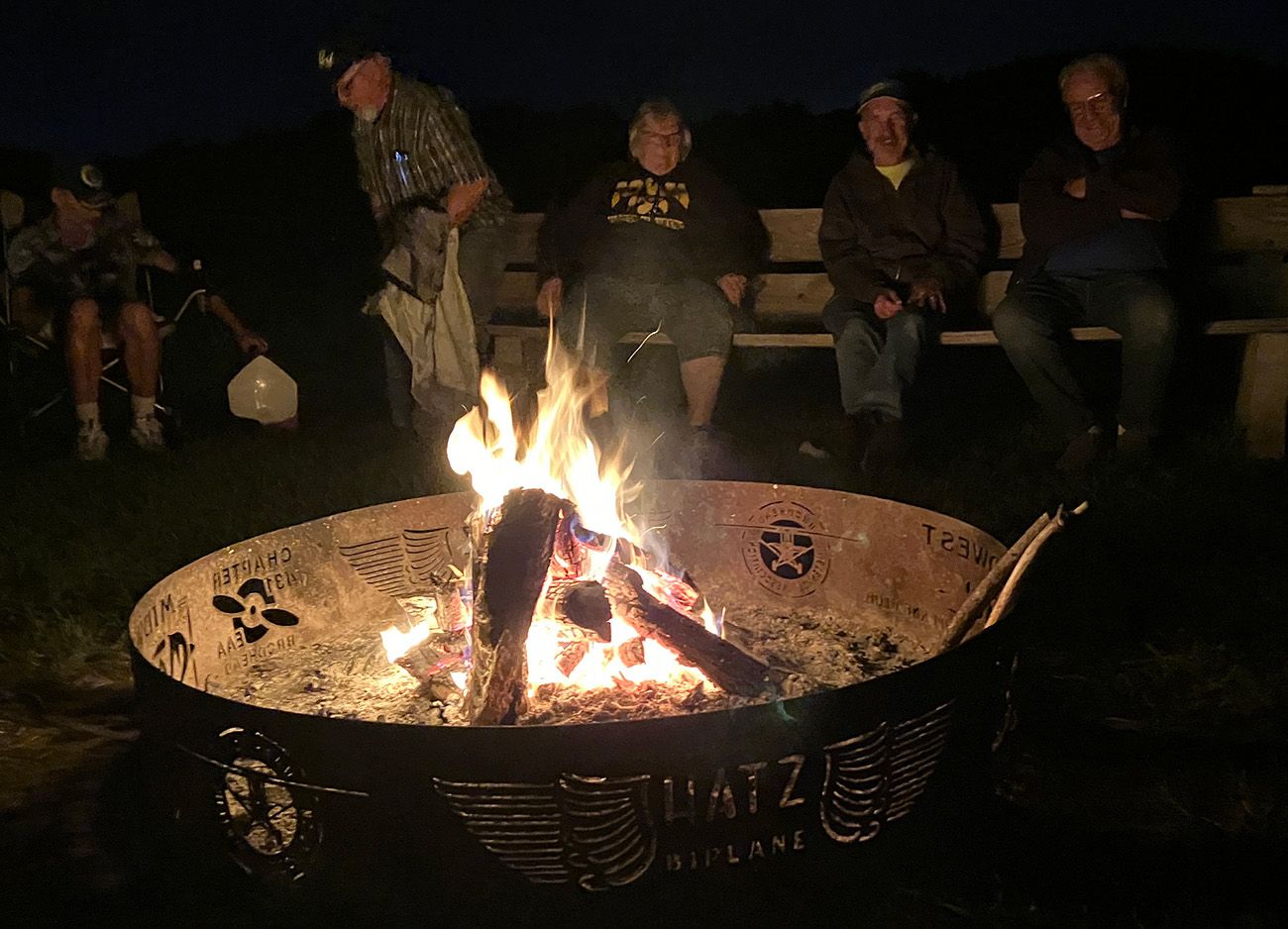 pilots gathered around a metal firepit at night. firepit has airplane motif cut out of sides.